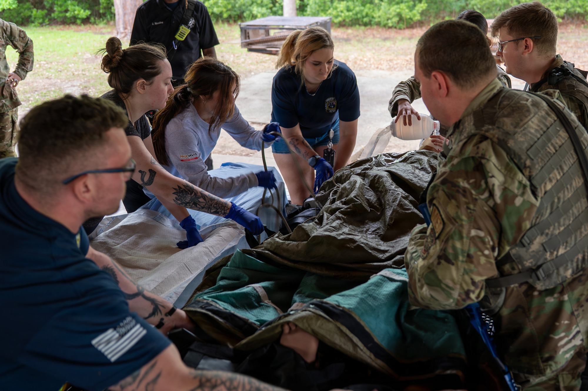 Five people stand around a mannequin covered in blankets performing various medical tasks.