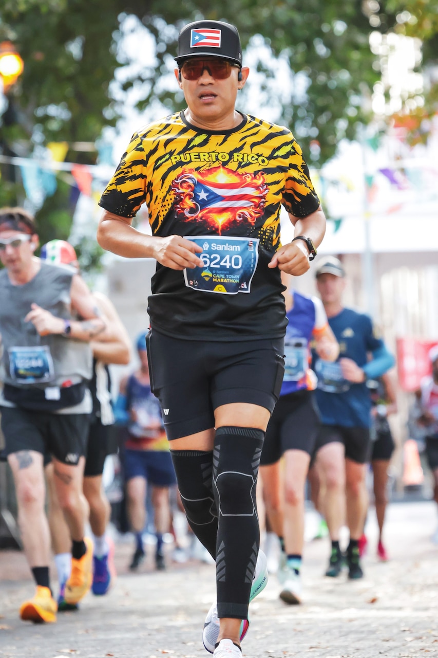 An airman wearing running gear jogs on a road during daytime, with other people in athletic attire also running in the background.