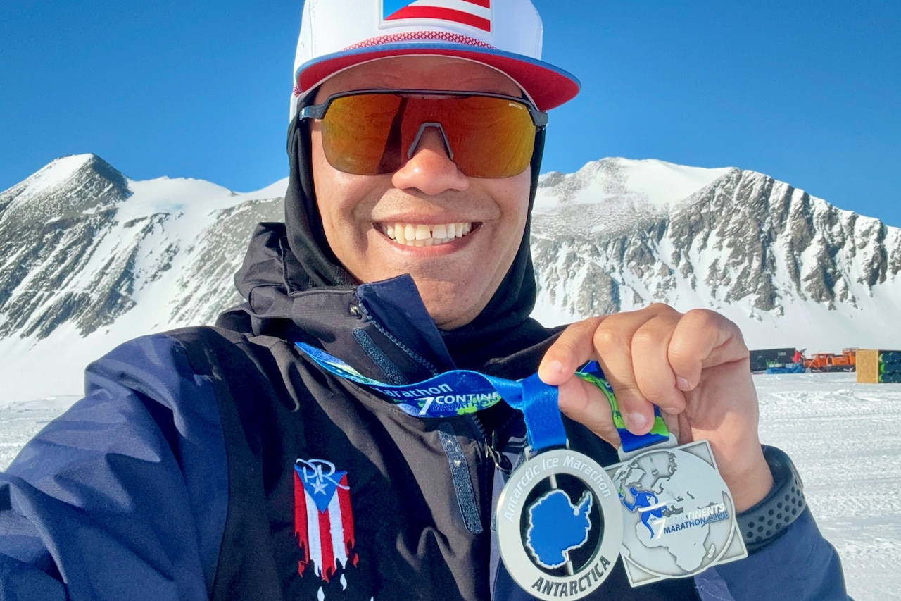 An airman wearing winter gear, a hat and sunglasses holds up two medals draped around his neck outdoors during daytime, with a snowy mountain in the background.