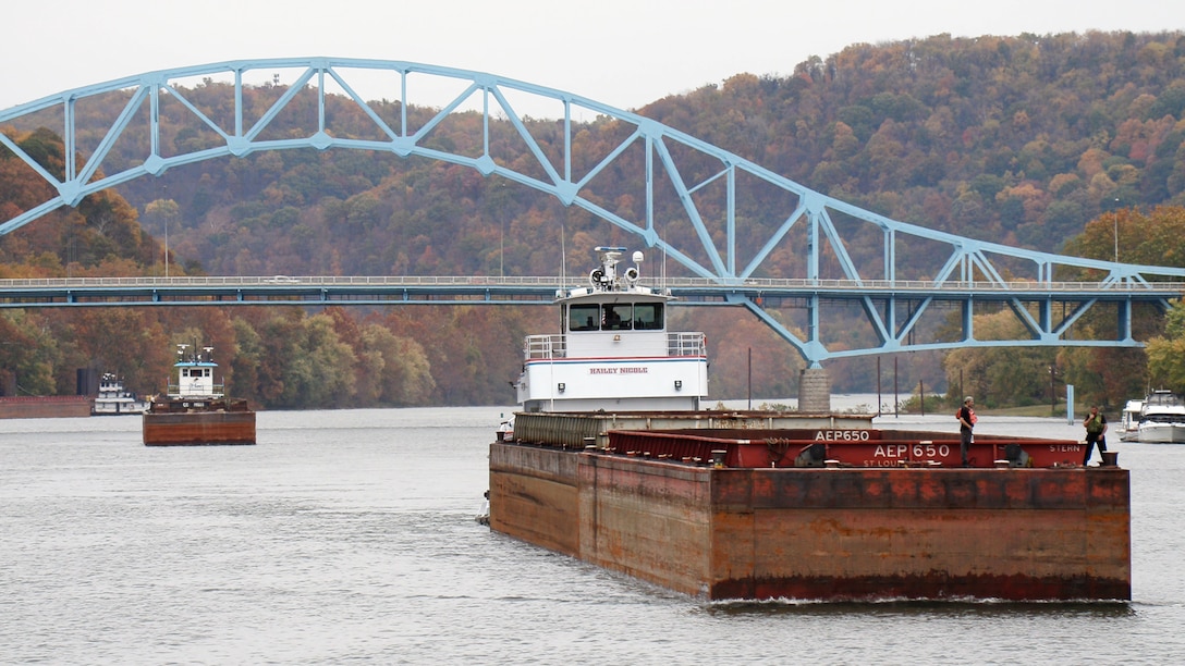 Two barges float down a river underneath a blue bridge in the fall, with colorful leaves in the background.