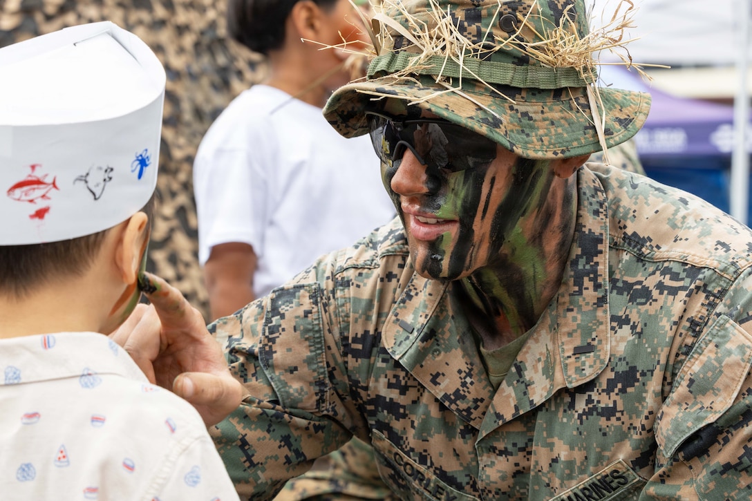 A Marine wearing camouflage on their face squats to paint it on a child’s face during the day.