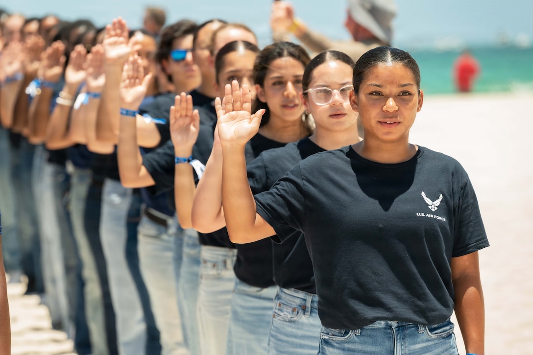 More than a dozen future service members in black shirts raise their right hands while standing in front of the ocean on a sunny day.