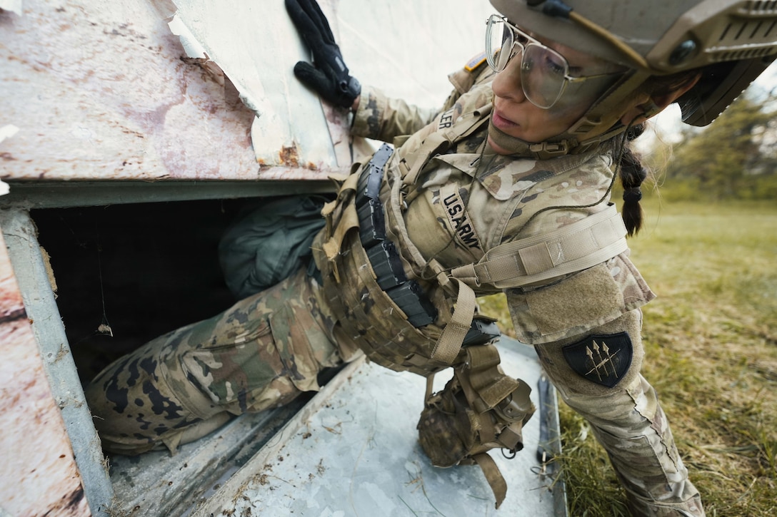 A soldier in tactical gear climbs into a square window during the day.