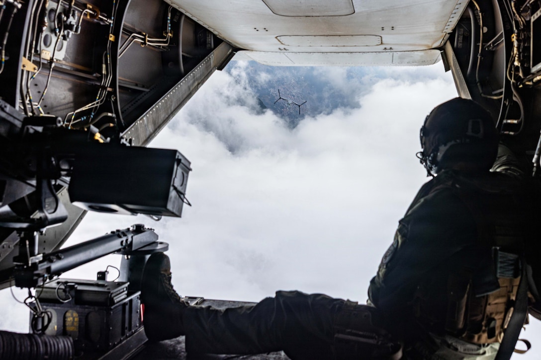 A Marine watches an aircraft fly in a cloudy sky while sitting in the open doorway of another aircraft.