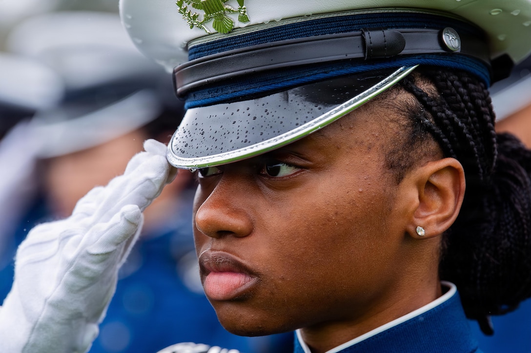 A cadet in a military dress uniform salutes, with other cadets in similar uniforms blurred in the background.