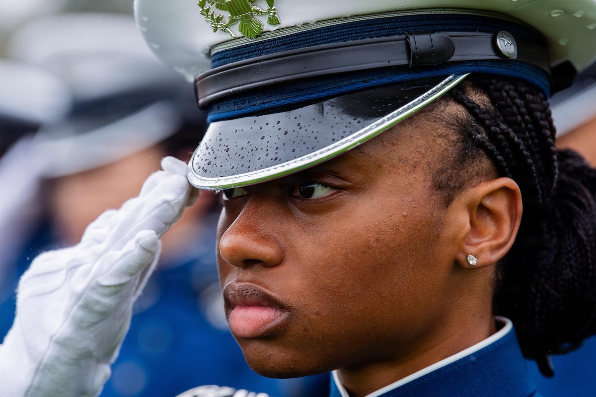 A cadet in a military dress uniform salutes, with other cadets in similar uniforms blurred in the background.