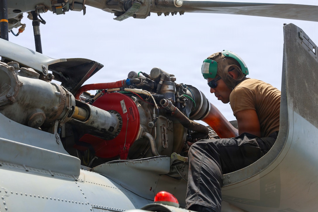 A sailor wearing a green helmet looks at machinery in a helicopter, with a light gray sky in the background.