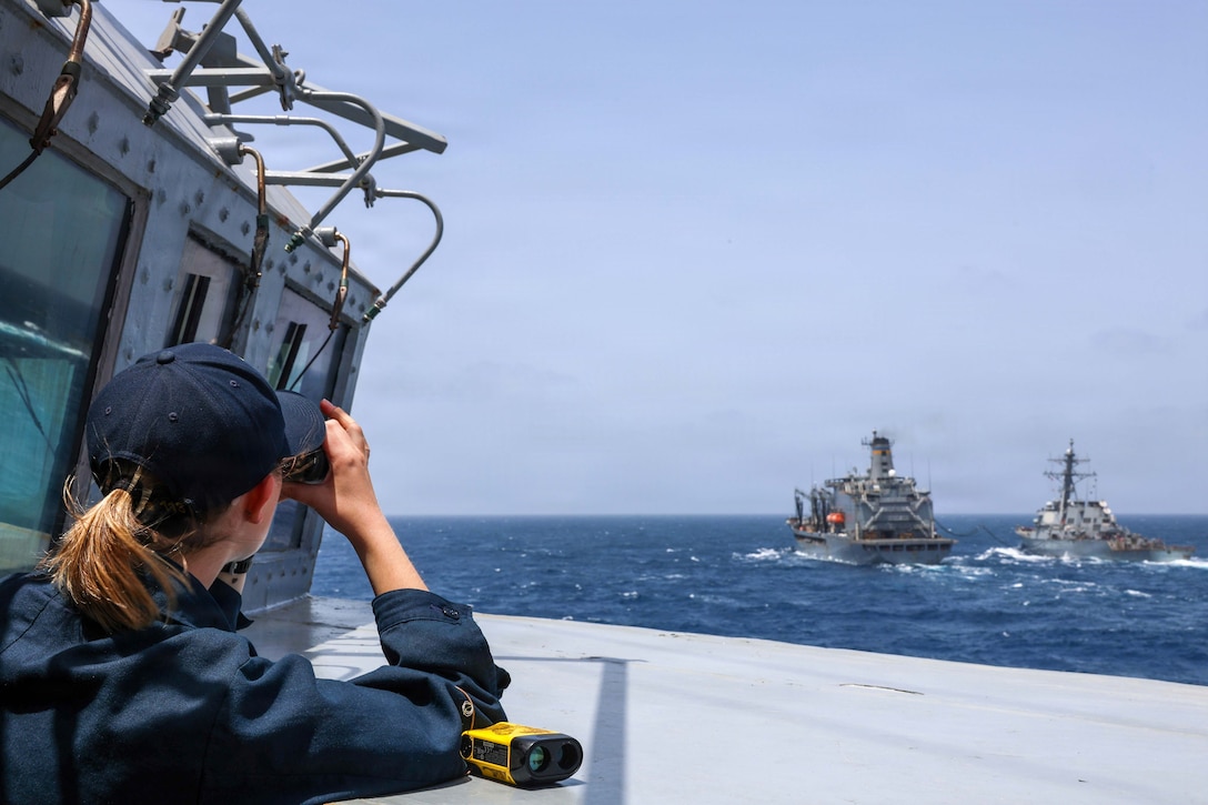 A sailor on a ship's deck looks out of a laser rangefinder toward two other ships, with water and the horizon in the background.