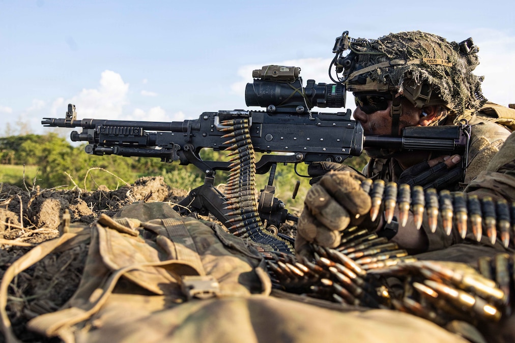 A soldier holds onto a gun that has ammunition draped over it and looks straight ahead while on the ground outdoors.