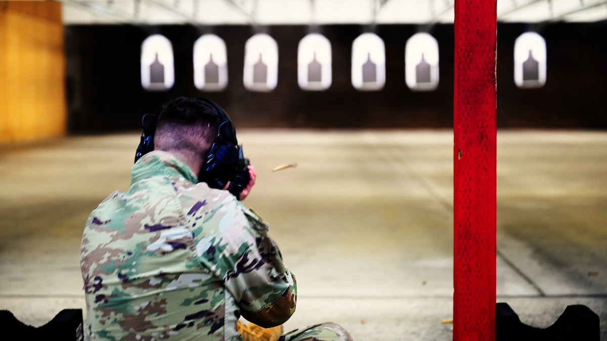 The back shoulders and head profile of an Airman shooting a weapon at a target downrange. A series of targets lined up, side by side, on the range can be seen in the background.