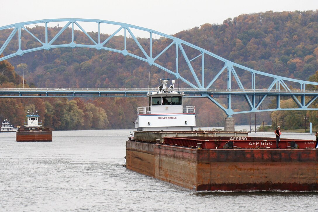 Two barges float underneath a light blue bridge on a river.