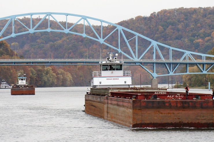 Photo of a barge on the Monongahela River with autumn color trees behind the bridge and towboat.