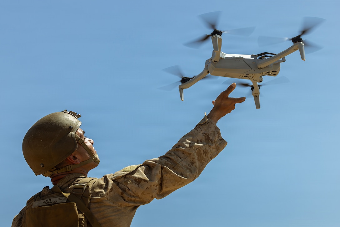 A Marine holds up their right arm while launching a small drone into blue sky.
