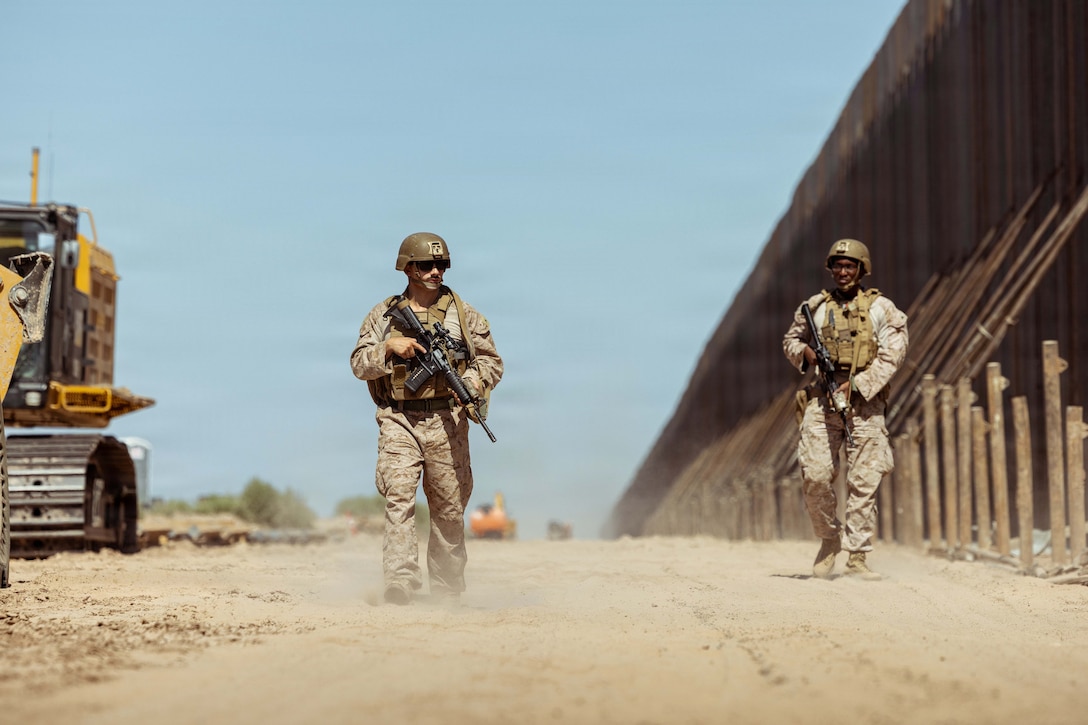 Two Marines walk on a dirt path holding machine guns, with a border wall on one side  of the path and construction equipment on the other side.