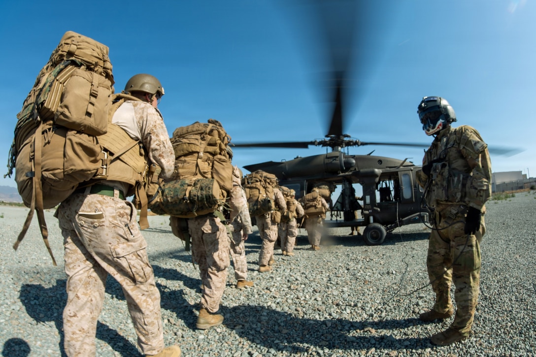 Marines stand in a line with backpacks to board a helicopter that is parked in front of them. Another Marine stands to the side watching them.