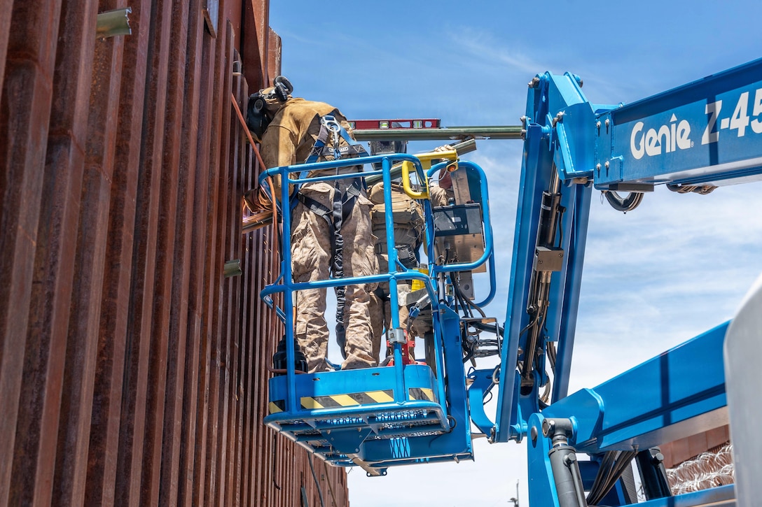 Marines in a blue lift work on a tall, metal fence during the day.