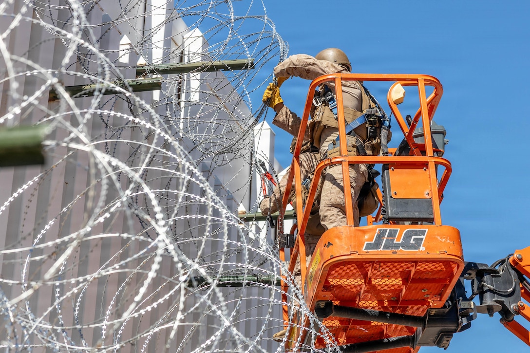 Marines in an orange lift work on barbed wire on the top of a metal fence against a blue sky.