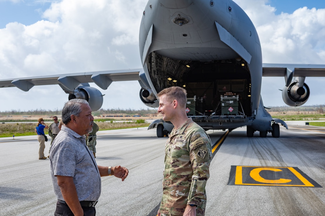 Two men, one in Army uniform, speak in front of the tail end of C-17 aircraft
