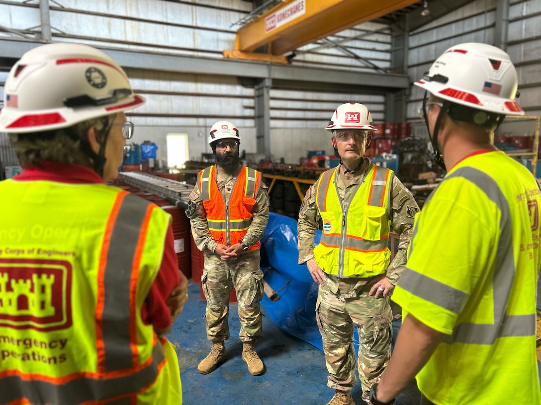 Two men in Army uniforms talk to two men in safety vests and hardhats inside of a power plant