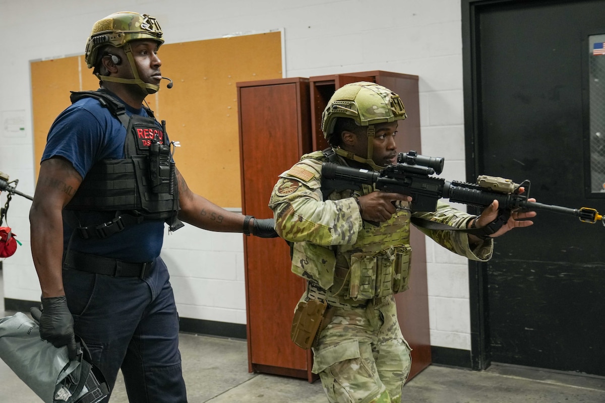 an airman points a gun forward as he leads an EMT through a hallway
