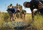 People in civilian clothing and military uniforms inspect munitions in a grassy area during daylight with a military vehicle in the background.