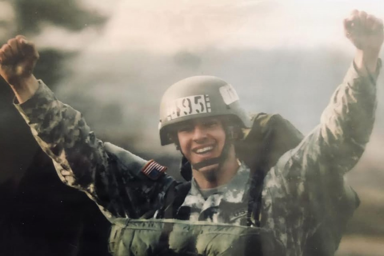 A man in a camouflage military uniform and helmet smiles and raises his fists in the air while wearing a parachute outside on the ground.