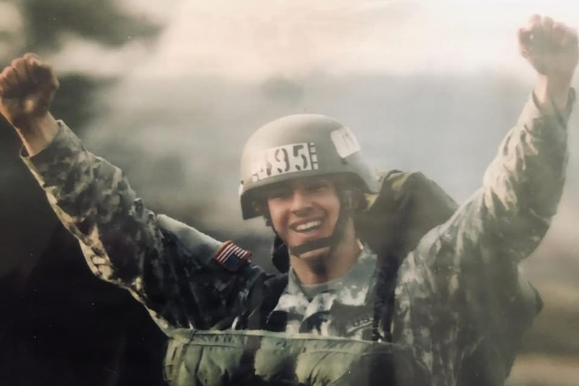 A man in a camouflage military uniform and helmet smiles and raises his fists in the air while wearing a parachute outside on the ground.