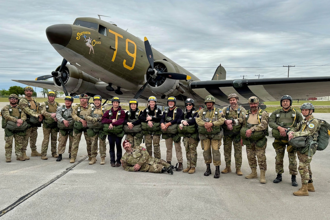 More than a dozen people in camouflage military uniforms pose in front of a World War II-era aircraft parked on a flight line. One person lies on the ground while the others stand.