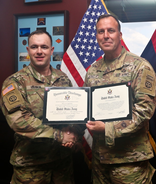 Two men in camouflage military uniforms hold a certificate and shake hands while posing for a photo indoors; an American flag is behind them.
