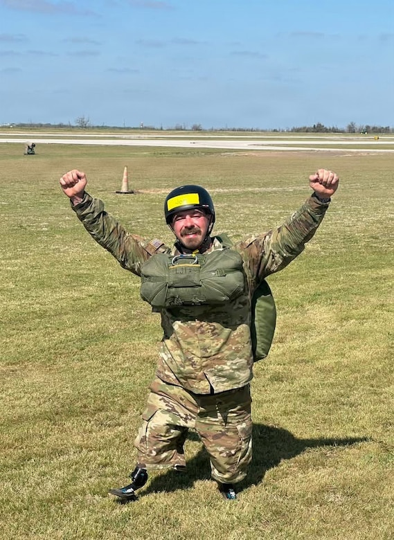 A man in a camouflage military uniform and helmet smiles and raises his fists in the air while wearing a parachute outside on the ground.