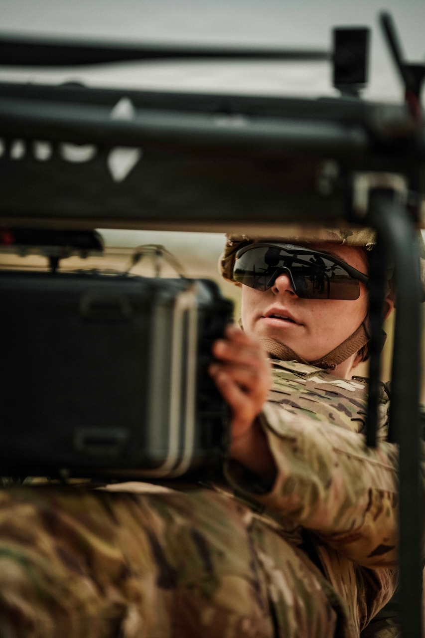 A person in a camouflage military uniform and sunglasses looks down at a screen outside while lying on their back.