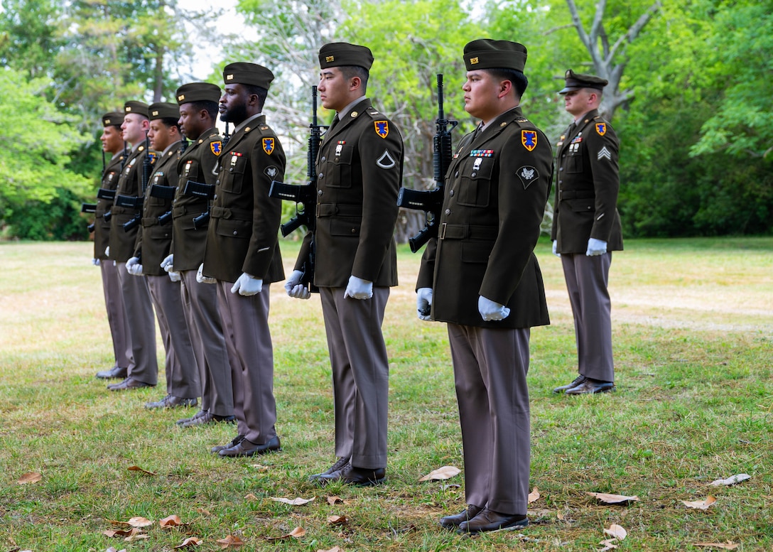 Photo of 7 soldiers in a line in dress uniform and white gloves holding rifles. There is one solider in dress uniform and white gloves standing behind the line.
