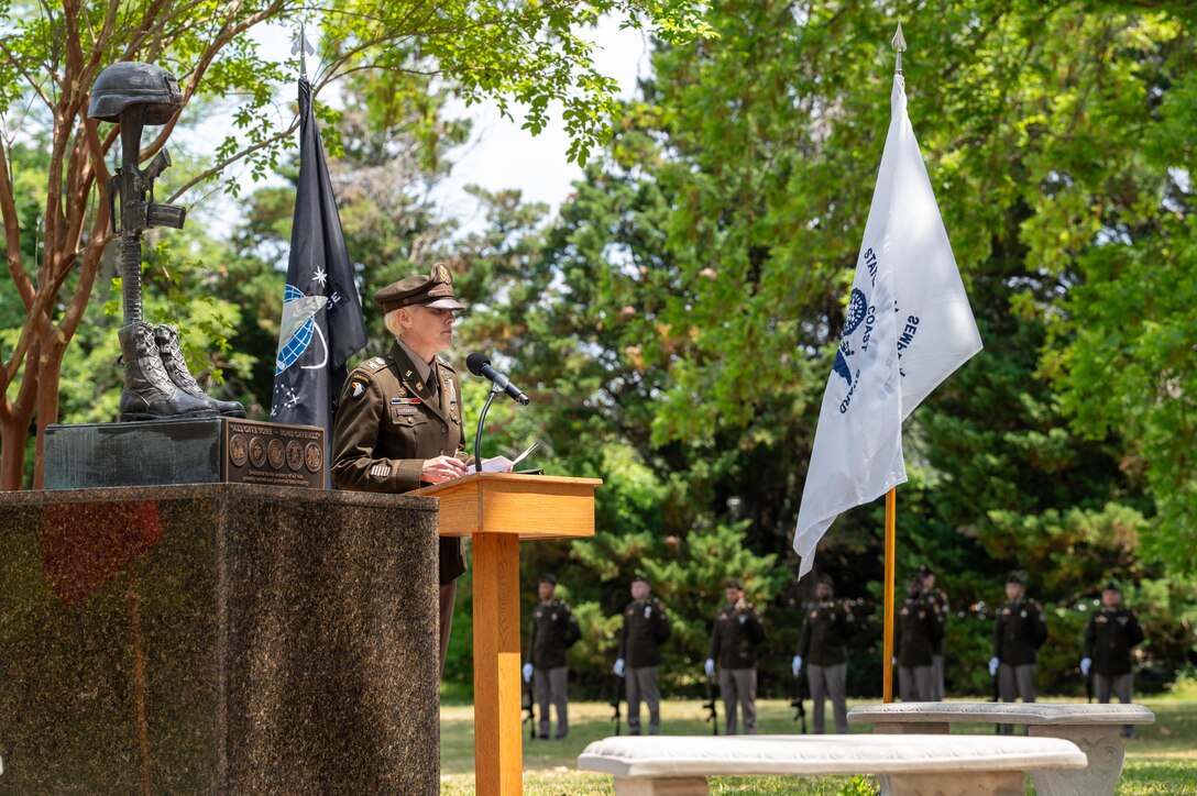 Photo of a woman in Army dress uniform speaking at a podium with flags in the background and to the right of a fallen solider statue.