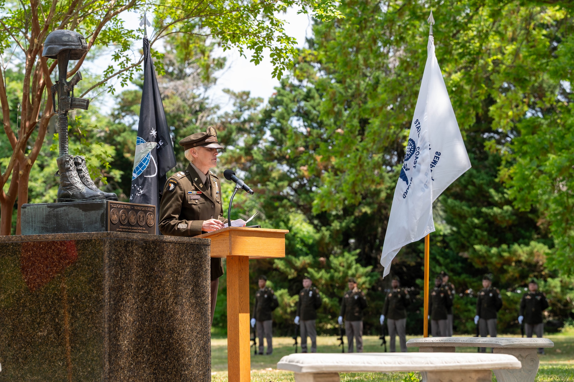 Photo of a woman in Army dress uniform speaking at a podium with flags in the background and to the right of a fallen solider statue.