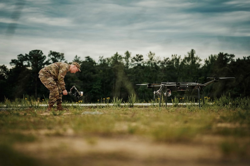 A man in a camouflage military uniform uses a device to take a photo of a military drone in a grassy field.