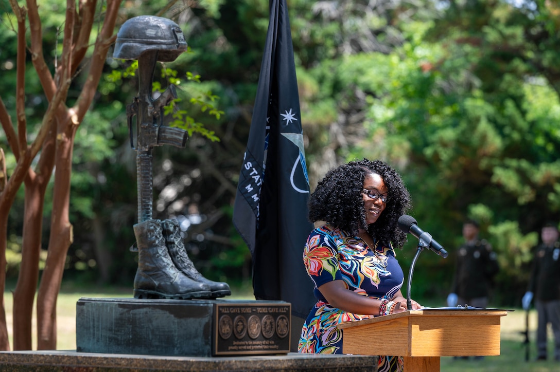 Photo of a woman speaking at a podium outdoors to the right of a fallen solider statue.