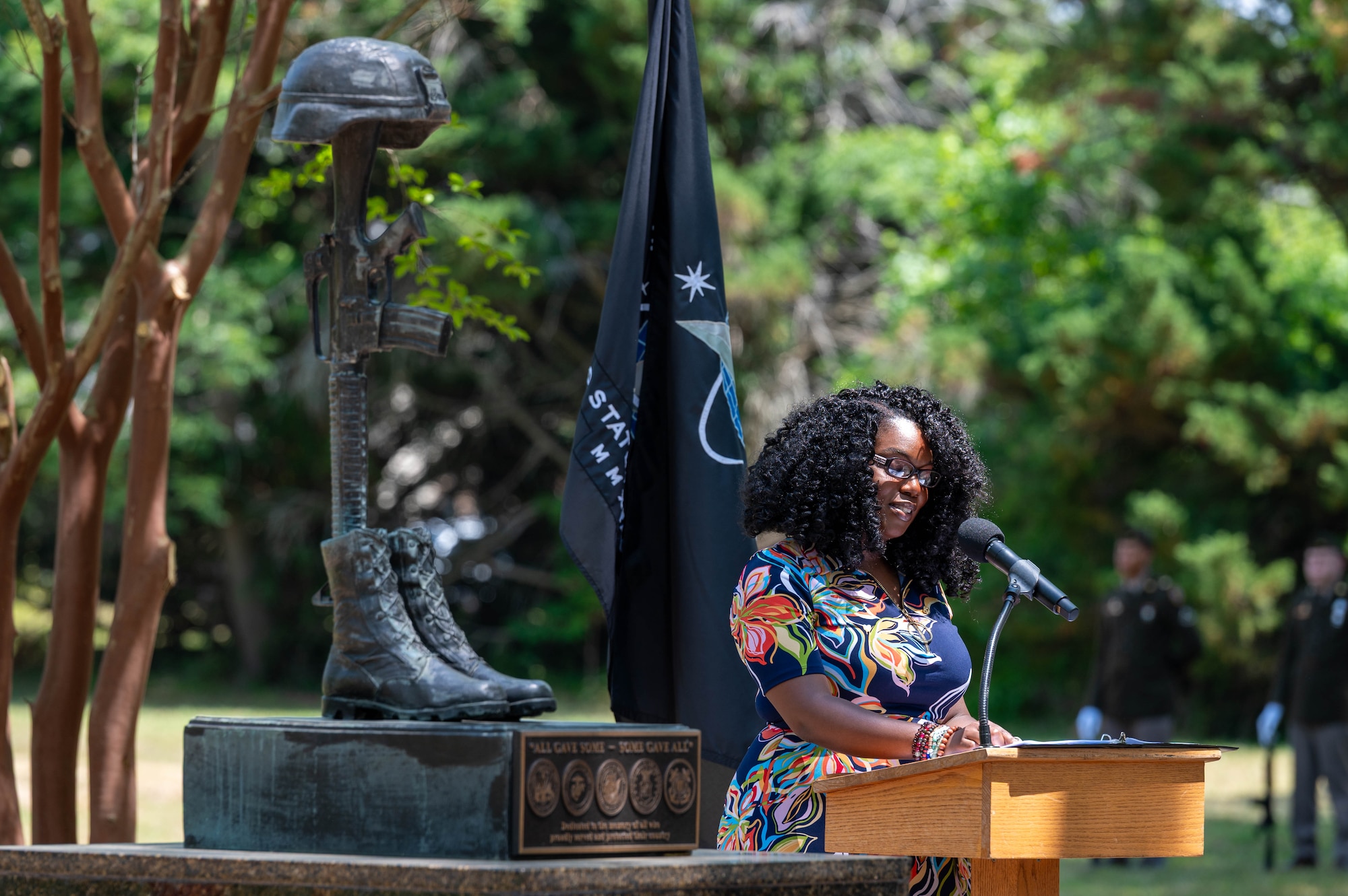 Photo of a woman speaking at a podium outdoors to the right of a fallen solider statue.