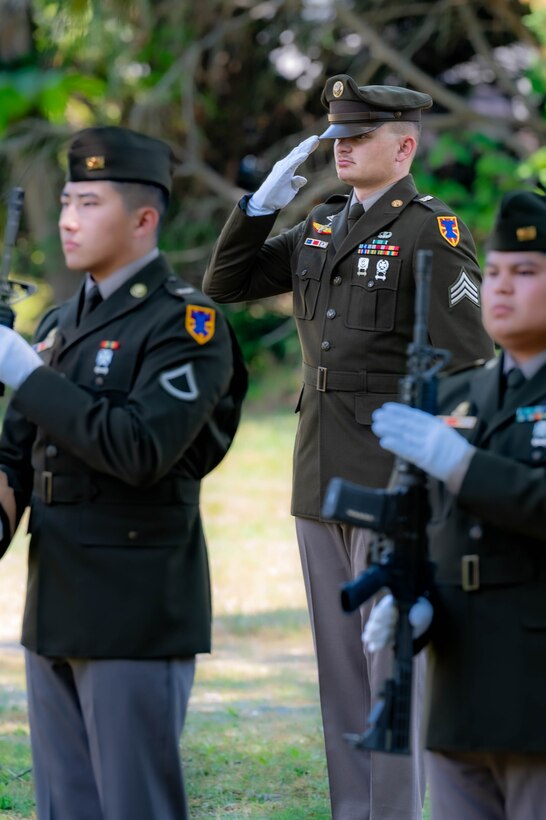 Photo of a soldier standing in the middle, but behind two other soldiers saluting. The solider on the right is holding a rifle. They are all in dress uniform, with white gloves.