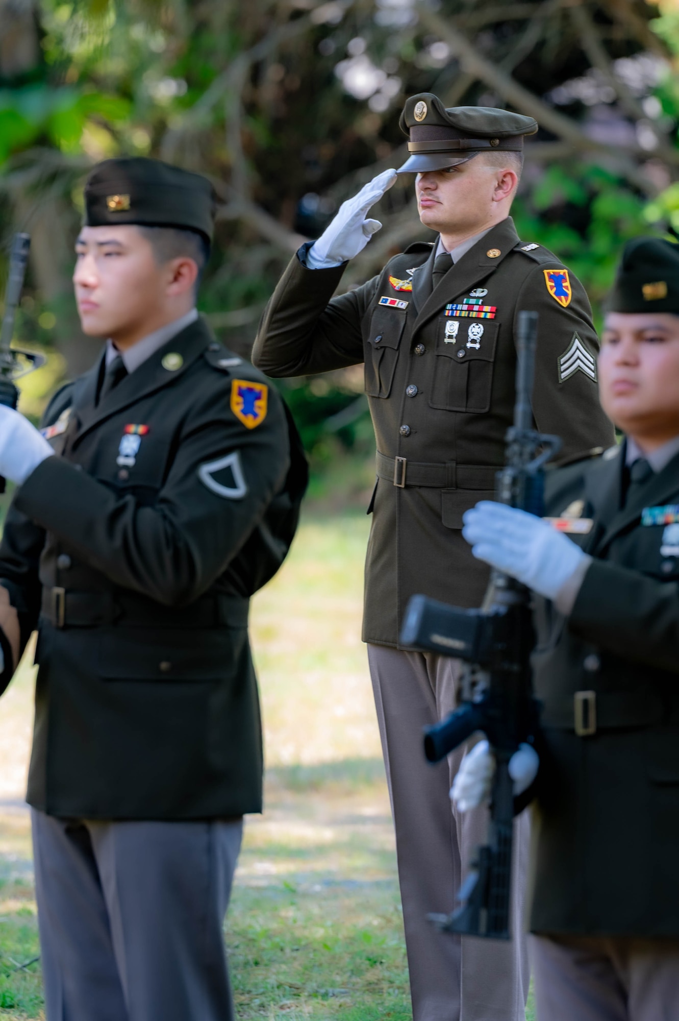 Photo of a soldier standing in the middle, but behind two other soldiers saluting. The solider on the right is holding a rifle. They are all in dress uniform, with white gloves.