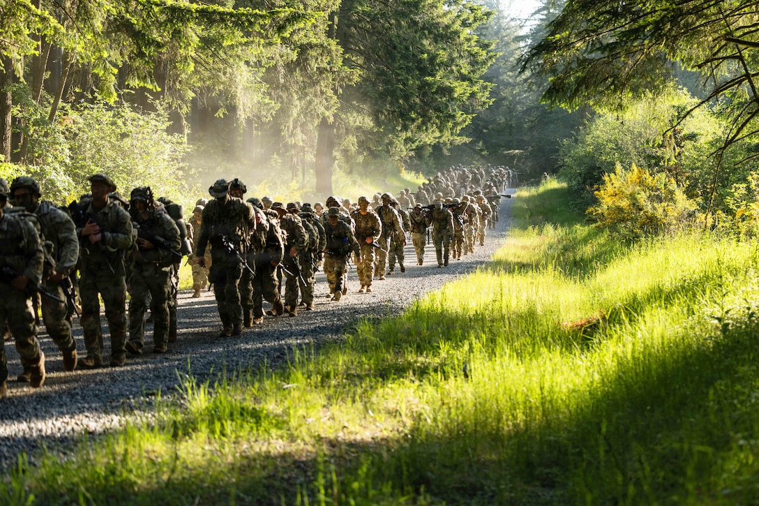 Dozens of soldiers in combat gear march through a shady, wooded area.
