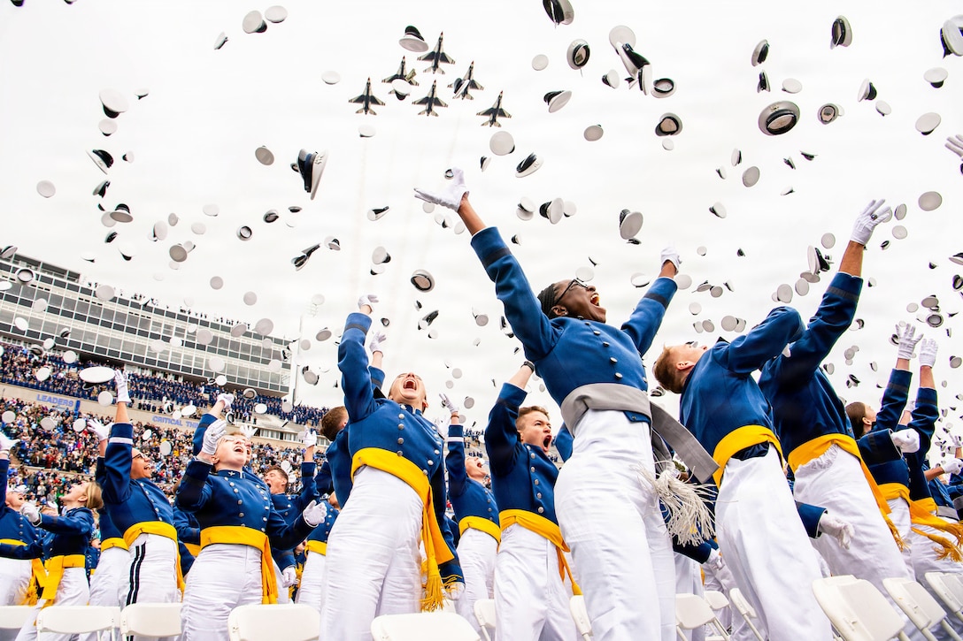 Dozens of graduates in ceremonial dress toss their hats into a blue sky as six aircraft fly in formation while loved ones watch from the stands.