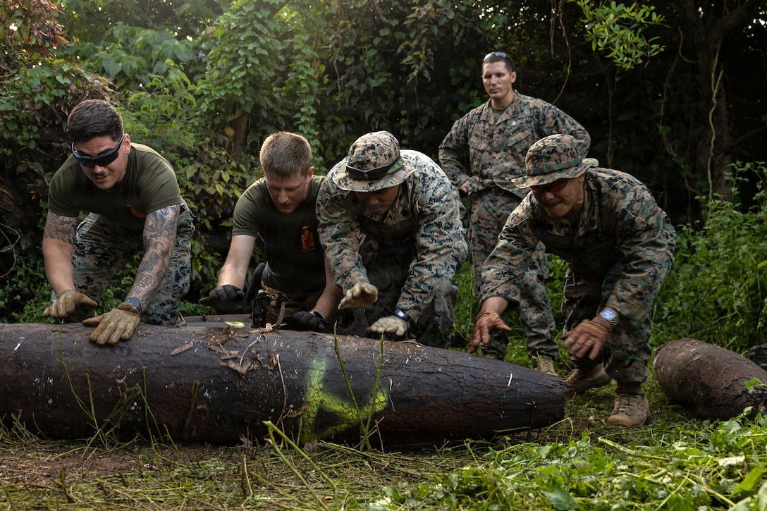 Marines roll a bomb dummy unit in a wooded area.
