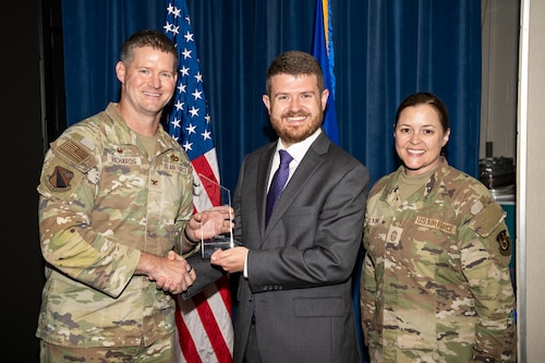 A man and woman, wearing military camouflage uniforms, stand on either side of a man, wearing a dark grey suit, posing for a group photo