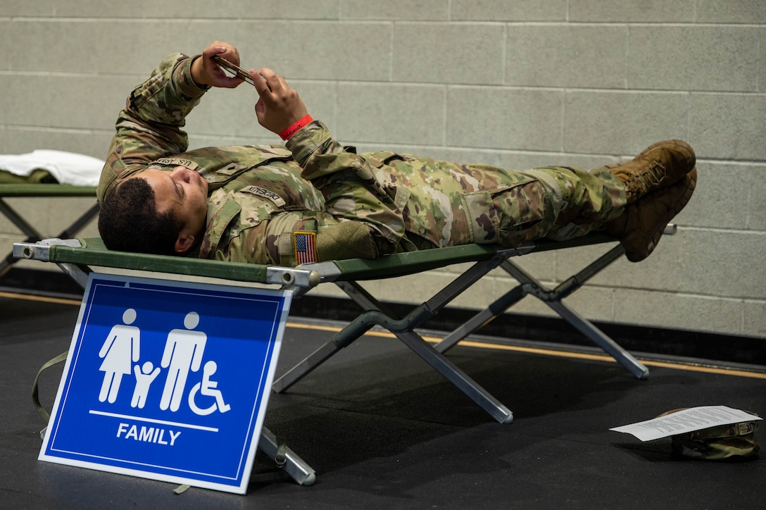 Photo of a solider laying on a cot, playing on his phone. There is a sign beneath the cot that says family.