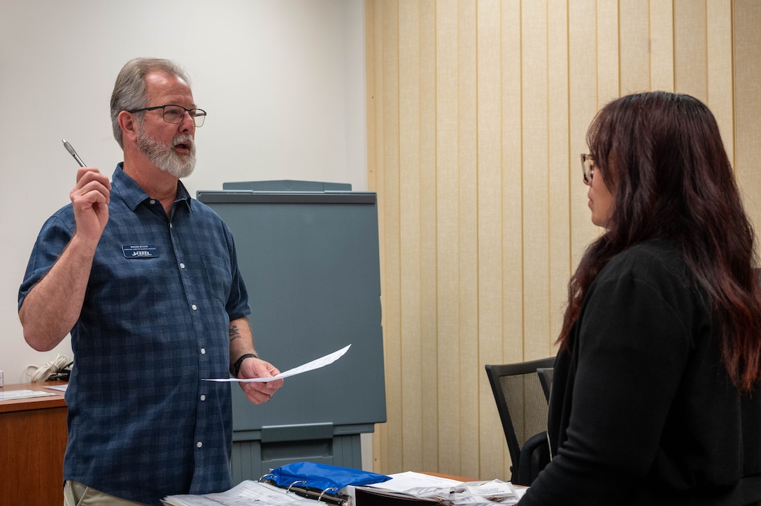 Photo shows a man on the left holding a pen in his left hand and paperwork in his right talking to a woman who is standing in front of him.