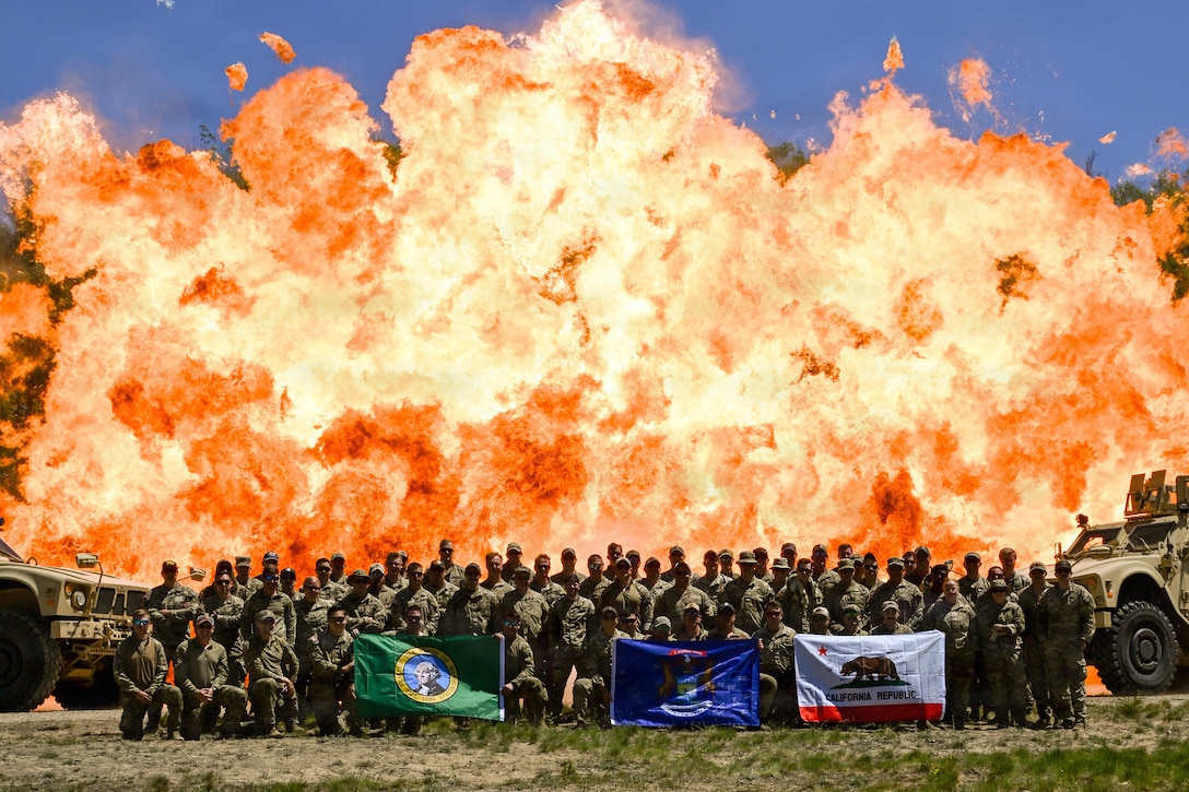 A large group of soldiers in camouflage pose for a photo, with flames rising high in the background.