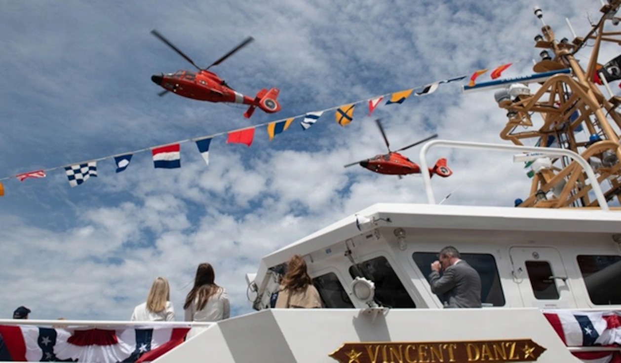 Two helicopters fly over a fast response cutter that is decorated with flags.