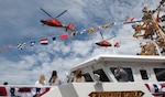 Two helicopters fly over a fast response cutter that is decorated with flags.