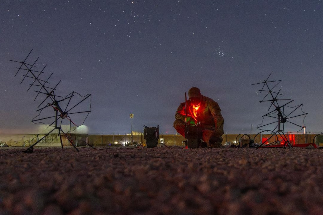 A soldier squats on the ground to look at a small piece of equipment with a red light, with an antenna to both sides and a starry sky above.