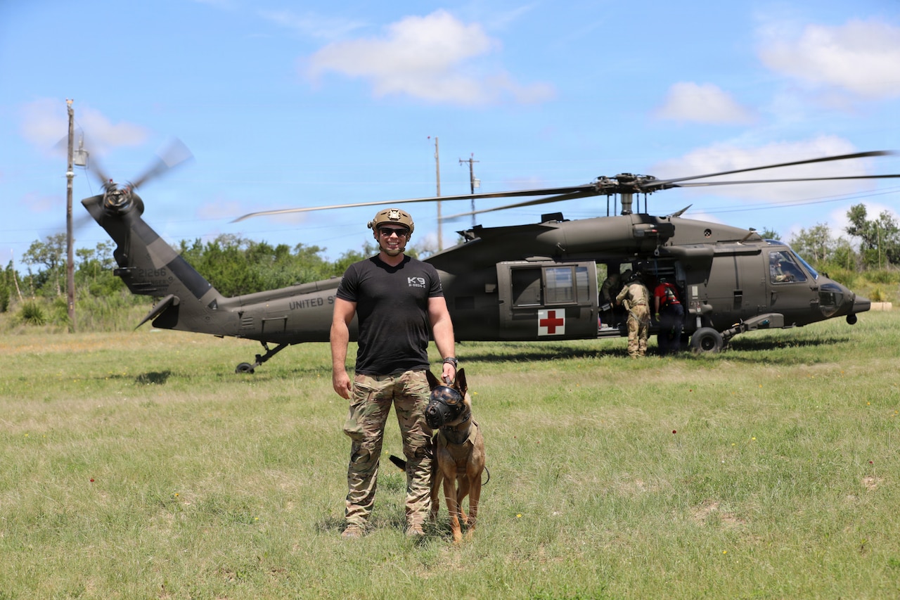 A man wearing a black T-shirt, camouflage military pants and helmet holds the collar of a dog as they pose for a photo while standing in front of a helicopter.